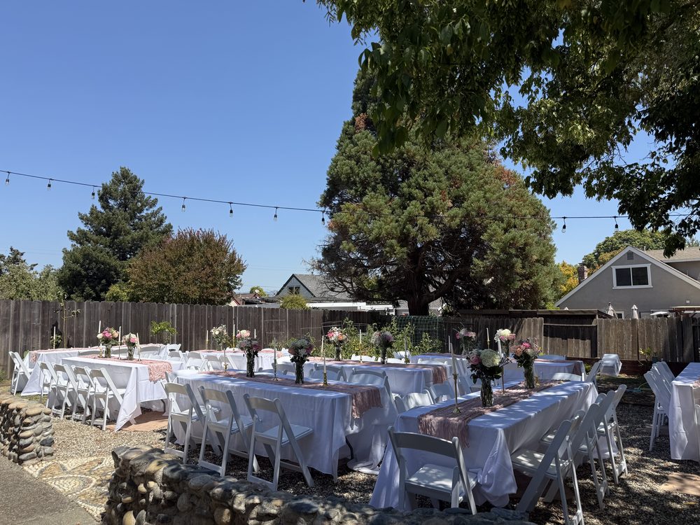 Long banquet tables with white linens, runners and white padded resin chairs set for an outdoor party